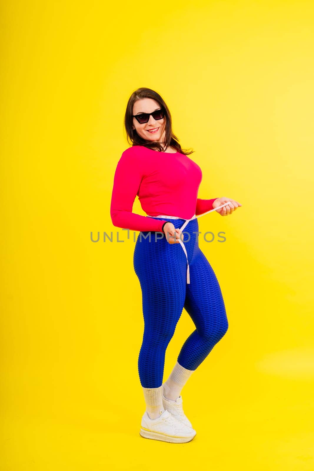 Pretty female with excess weight in a sporty top measuring waist over white yellow background