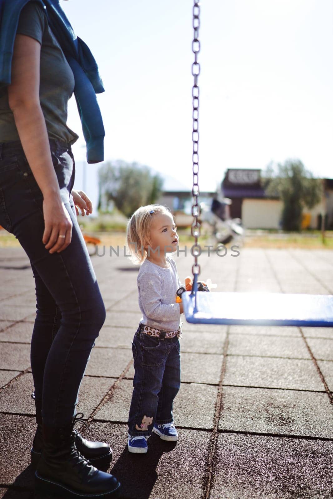 Mom stands near the little girl next to the chain swing on the playground. Cropped. High quality photo