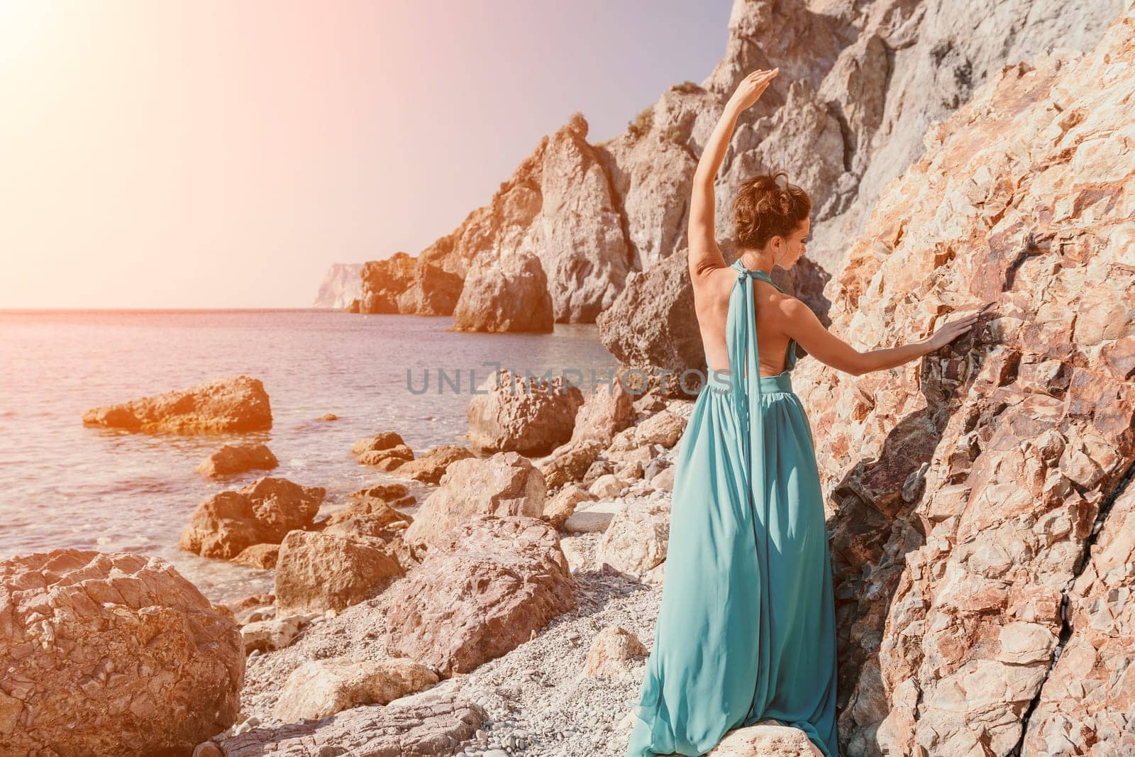 Side view a Young beautiful sensual woman in a red long dress posing on a rock high above the sea during sunrise. Girl on the nature on blue sky background. Fashion photo.