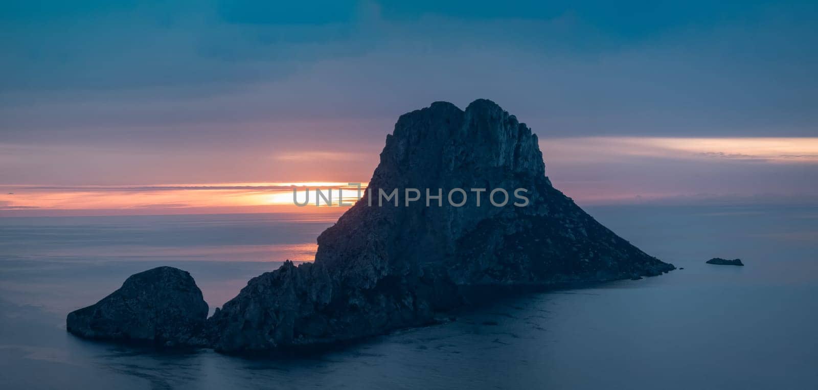Serene sunset at Es Vedra in Ibiza with the sun descending behind the iconic rock in a calm sea.