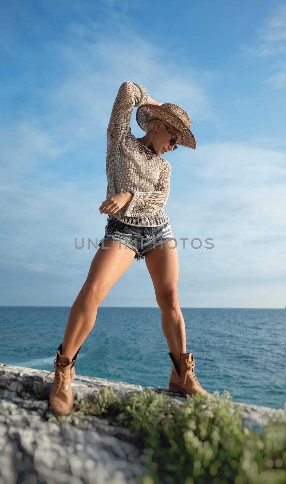 sexy girl in stylish fashionable clothes, cowboy hat and cool boots, posing freely on white rocks against the background of the sea