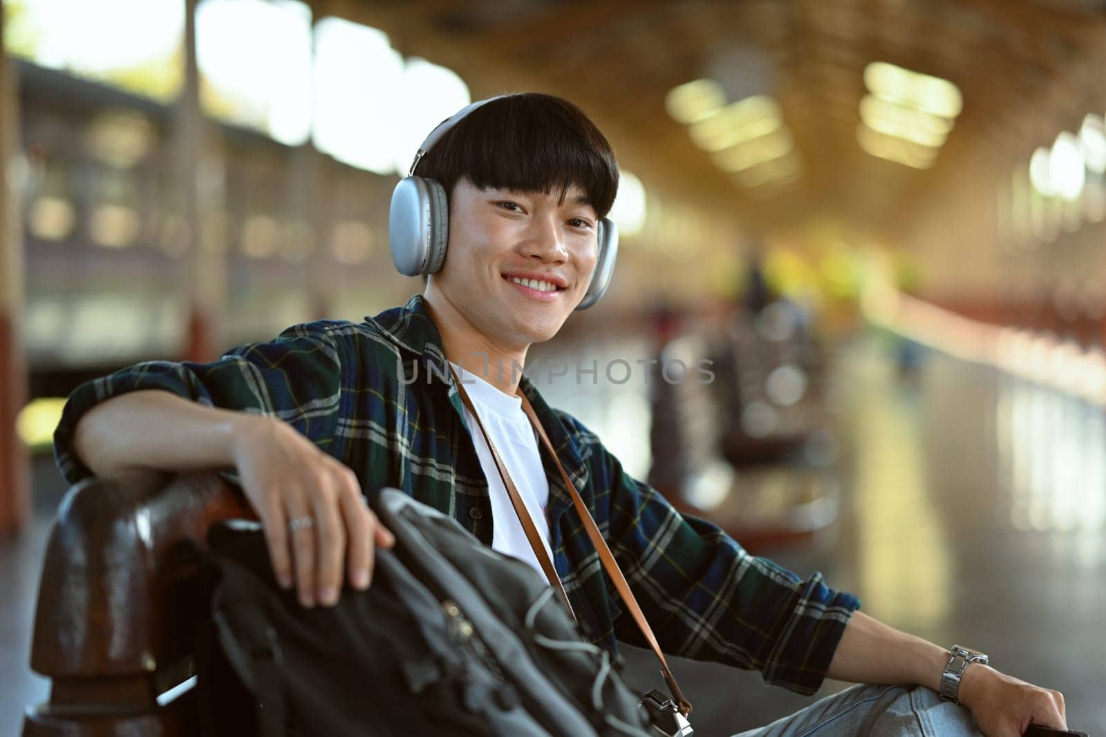 Portrait of smiling Asian man tourist in headphone waiting train at railroad station platform by prathanchorruangsak