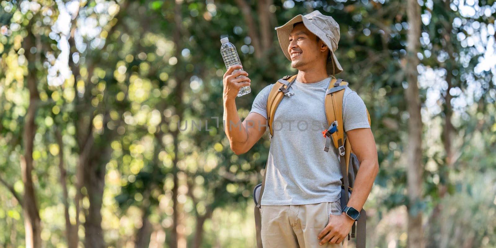 Young man backpacker traveling alone in forest. Attractive male traveler drinking water while walking in nature wood during holiday vacation trip by itchaznong
