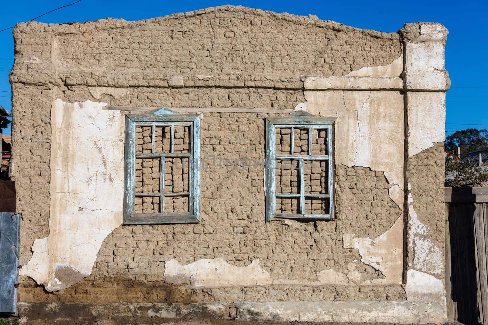 old house adobe air brick wall with two embedded wooden window frames and peeled off plaster leftovers - flat texture.