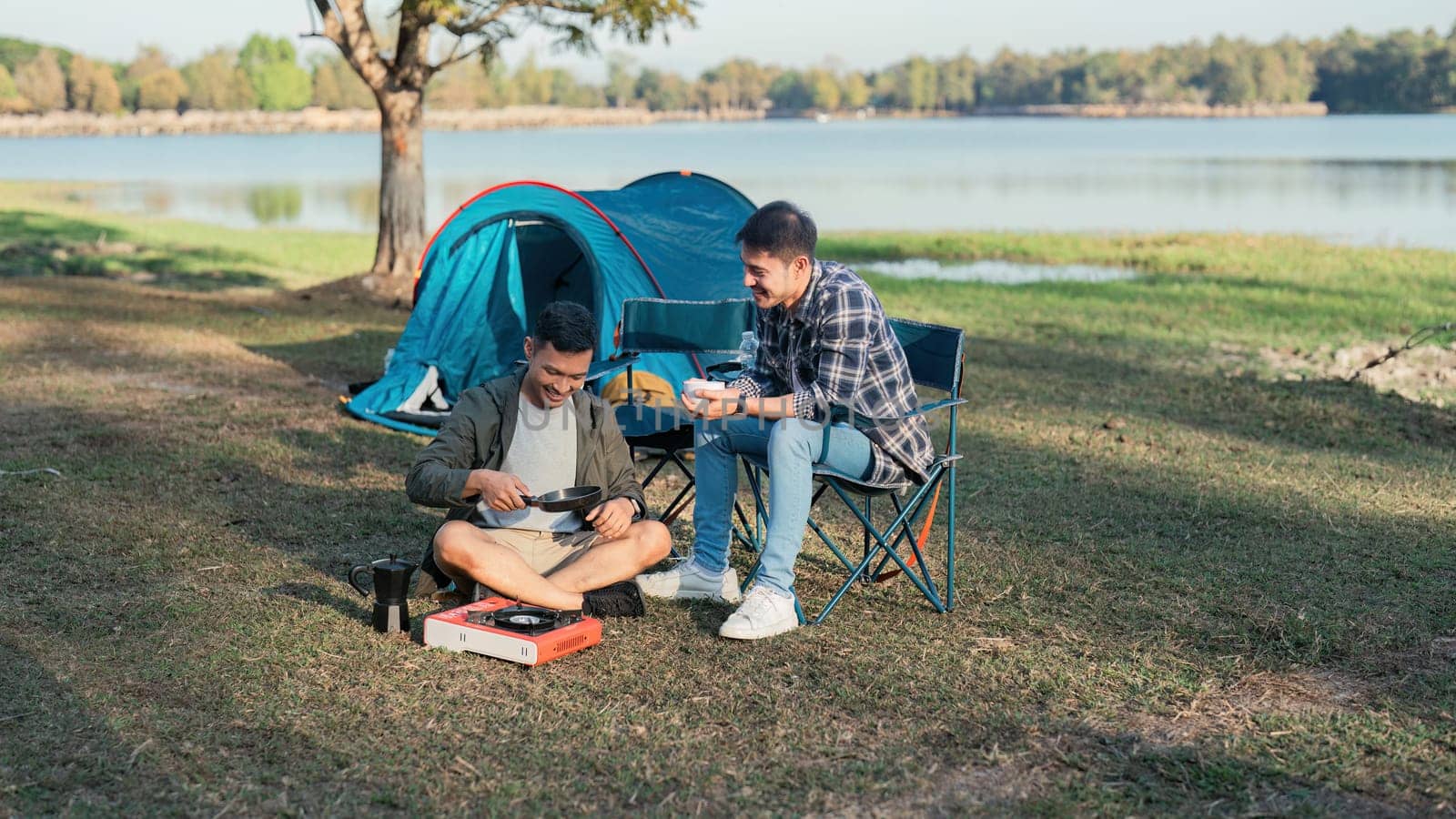 Gay LGBTQIA couple sitting on picnic chair drinking tea and making food while camping on vacation holiday by itchaznong