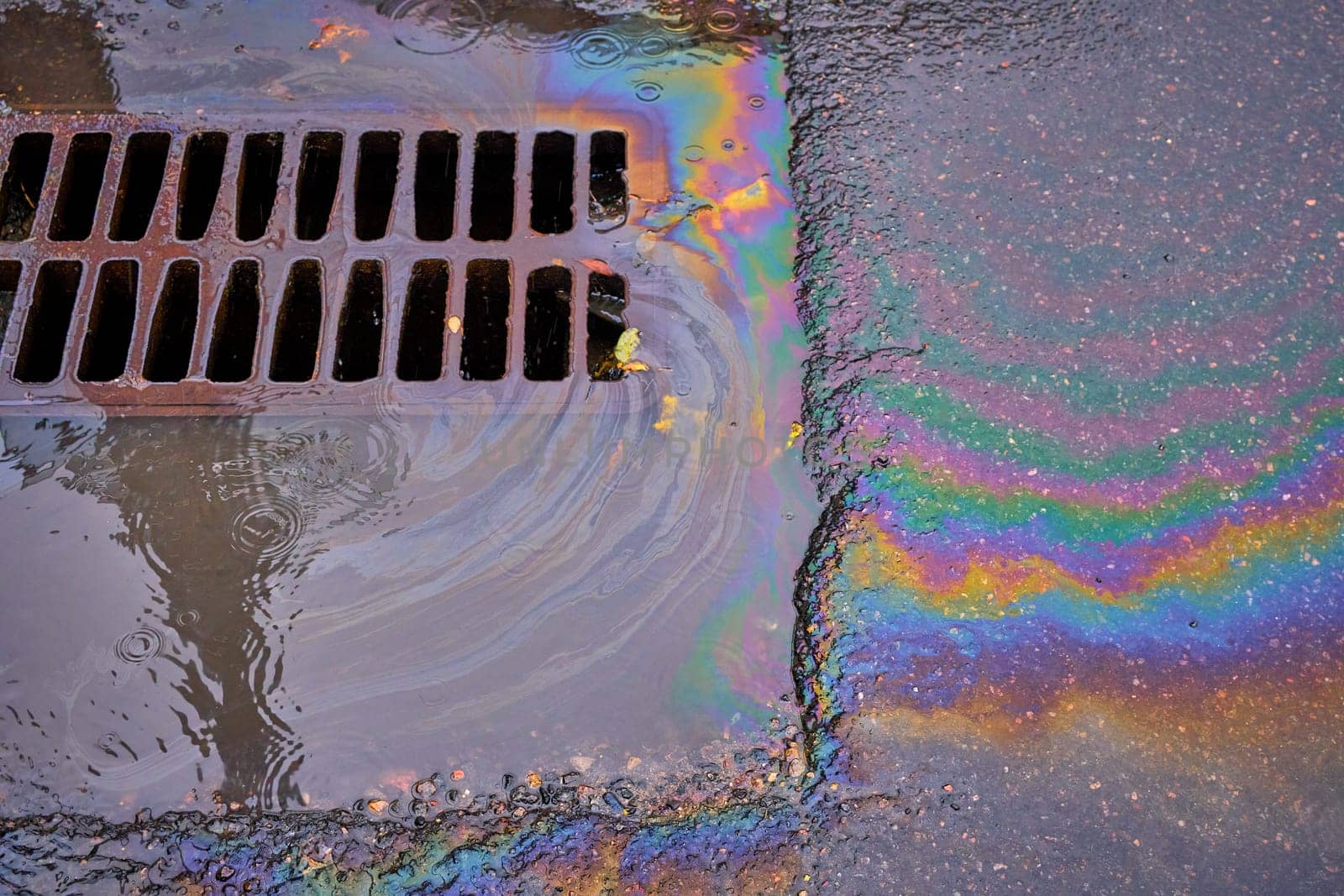 An oil slick against the backdrop of an asphalt road flows into a storm drain through a grate. Environmental problems of water pollution.