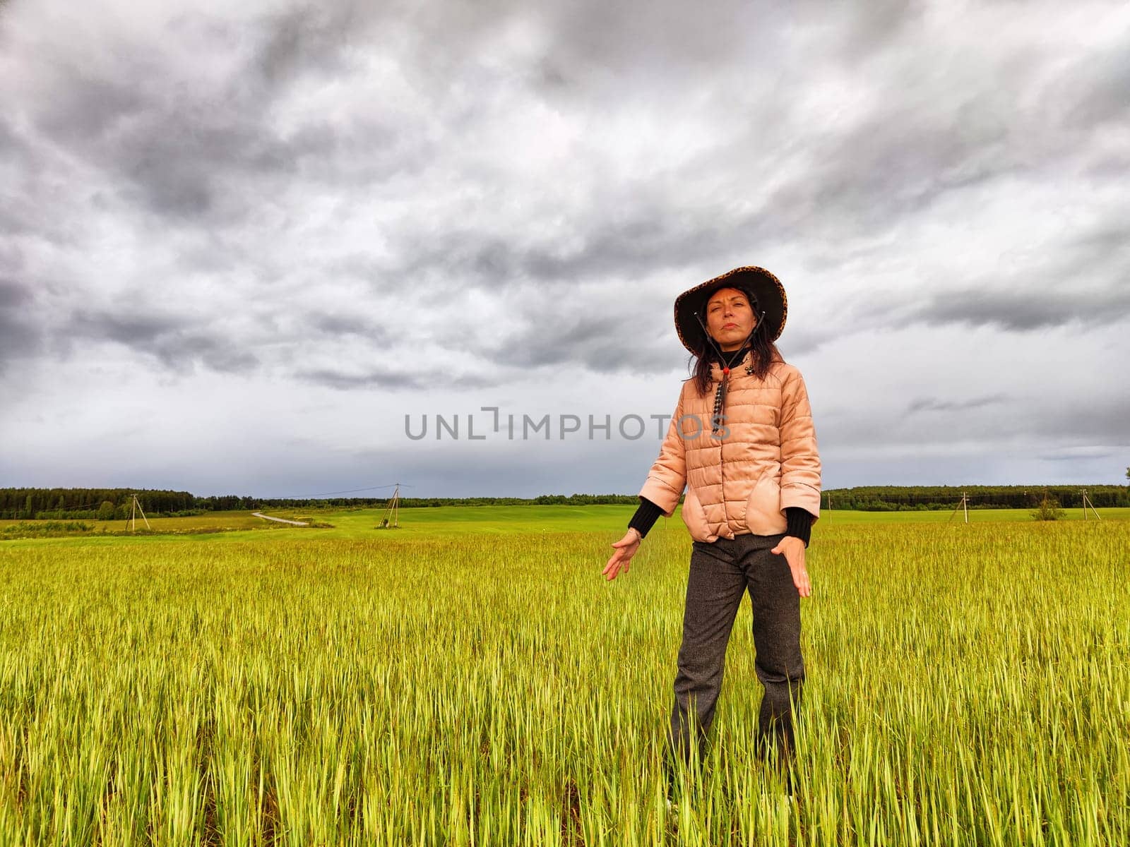 An adult girl looking like a cowboy in a hat in a field and with a stormy sky with clouds takes pictures of rainbow and takes selfie in the rain. Woman having fun outdoors on rural and rustic nature