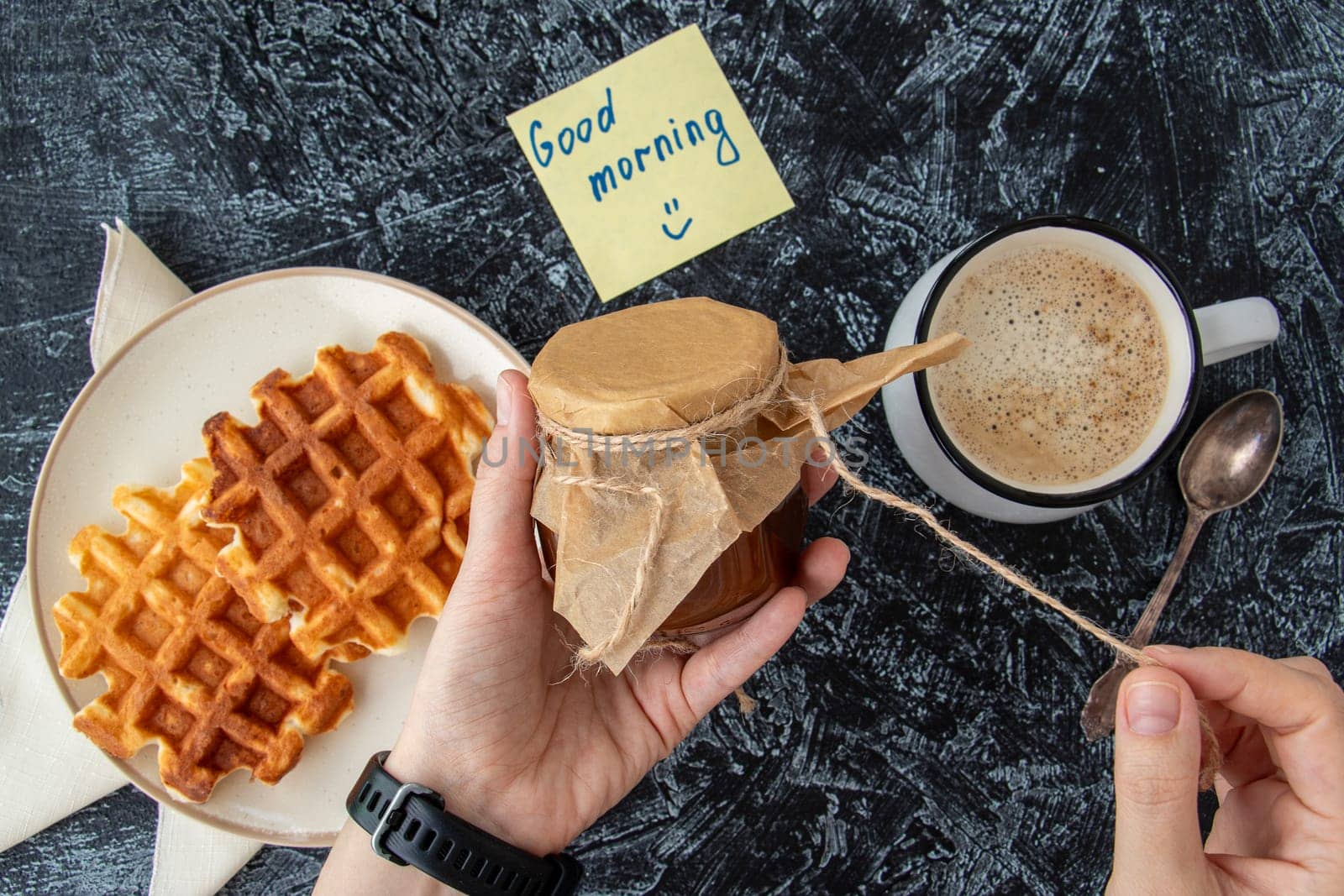 Top view of woman's hands holding a jar of homemade jam next to a cup of coffee and homemade Viennese waffles on a black textured background and a note Good morning. breakfast flat lay