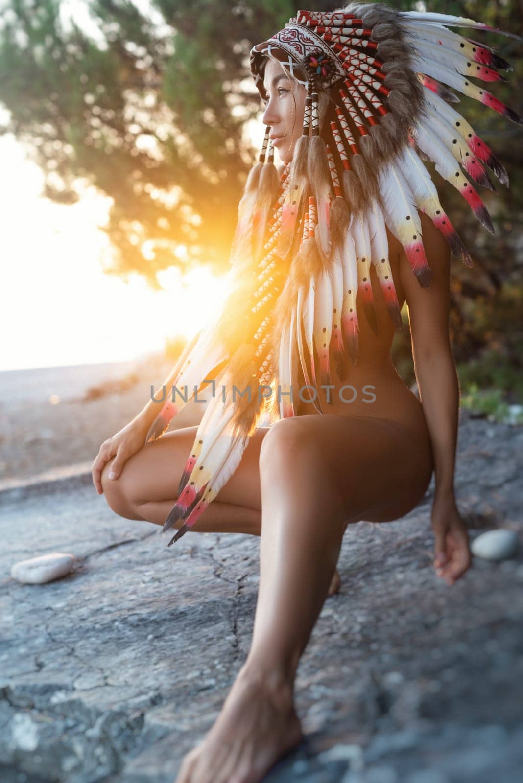 Naked sexy girl in Native American headdresses on the background of nature in the beautiful sunset light