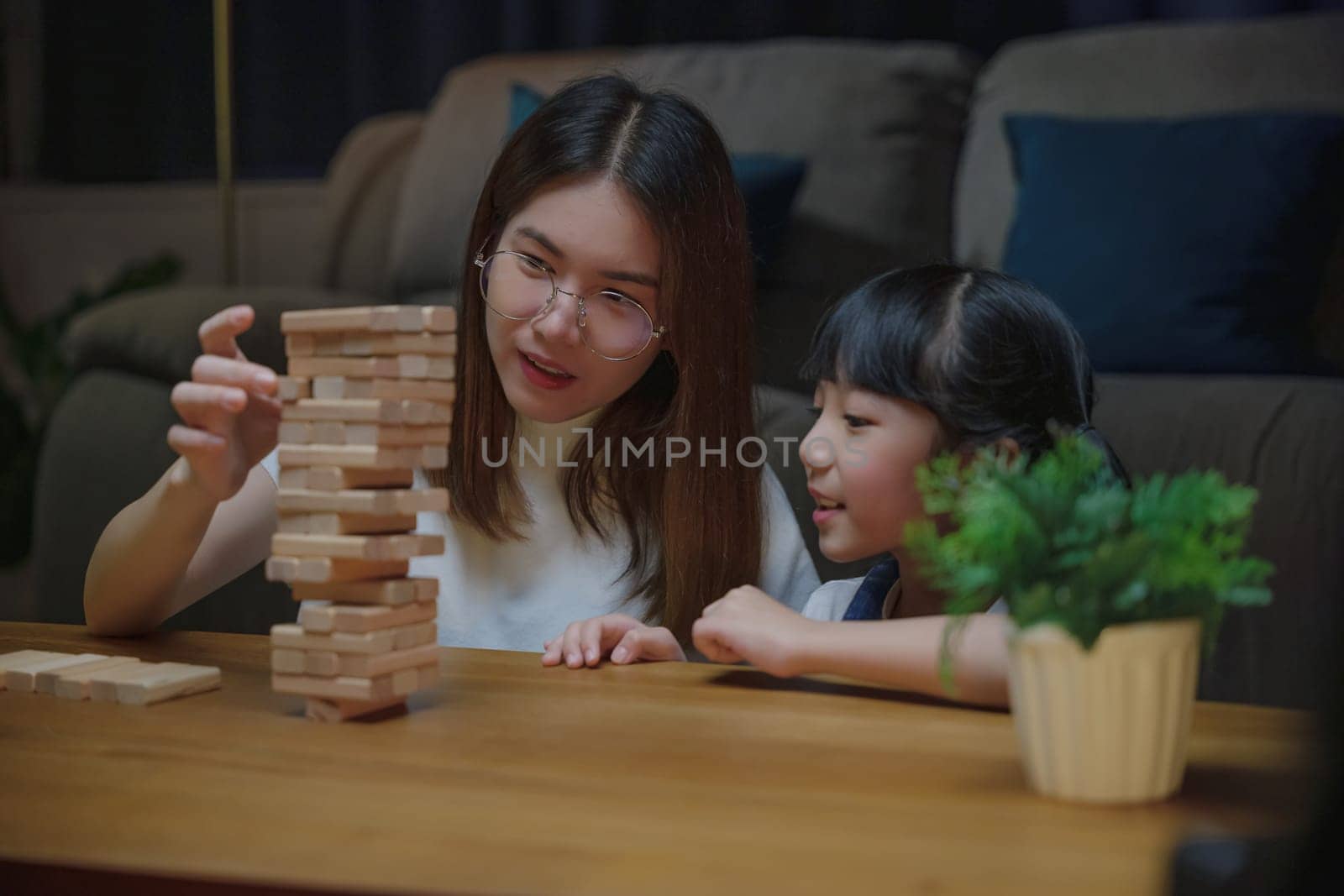 Asian young mother playing game in wood block with her little daughter in home living room at night time, Smiling woman help teach child play build constructor of wooden blocks, education