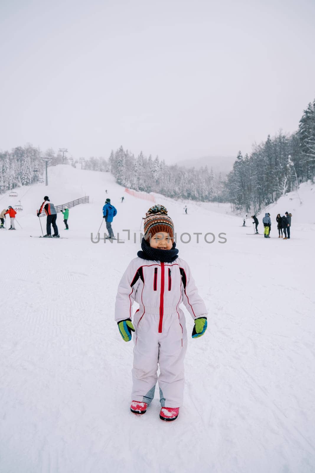 Little smiling girl stands on the ski slope against the backdrop of skiers. High quality photo