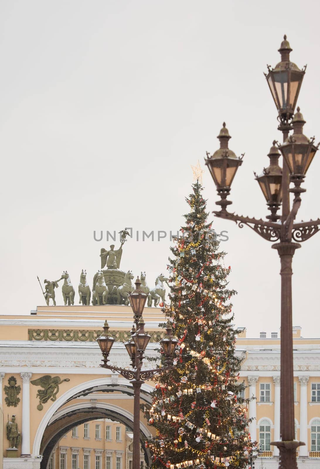 The main Christmas tree shimmers with lights of decorations through vintage lamppost, the central Palace square of the city decorated for the celebration during the snowfall, Arch of the General Staff. High quality photo