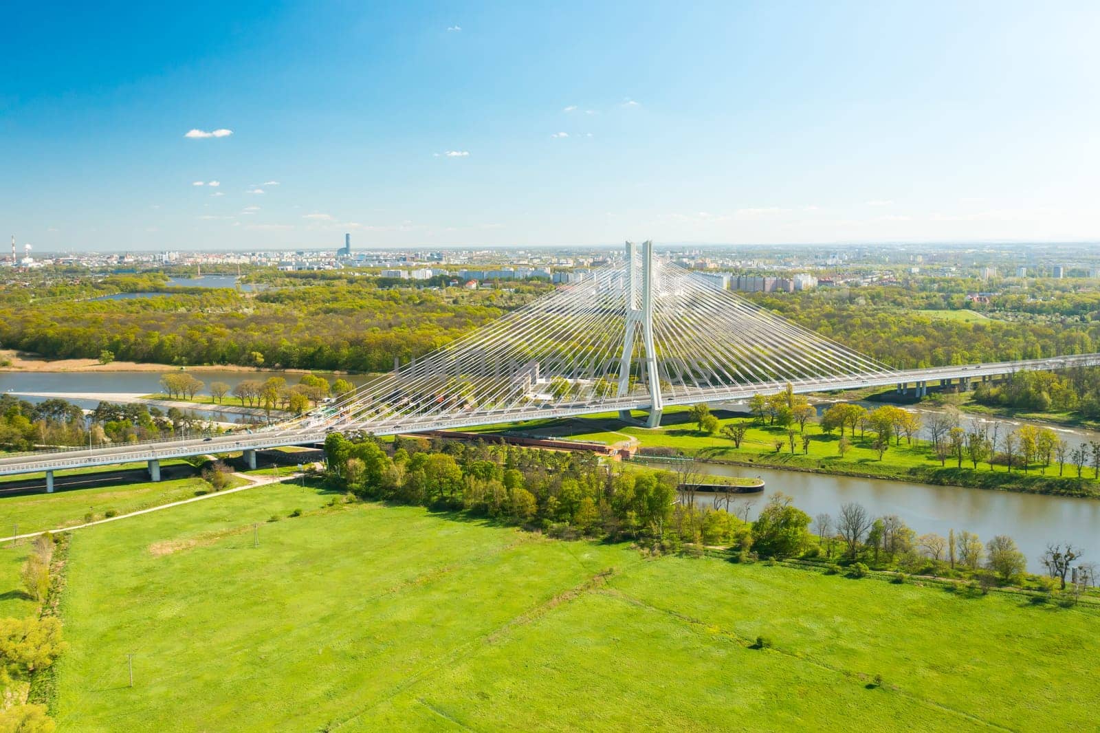 Cars drive on cable-stayed Redzinski Bridge over river flowing near scenic Wroclaw. Pylon bridge surrounded by lush green forests aerial motion along bridge
