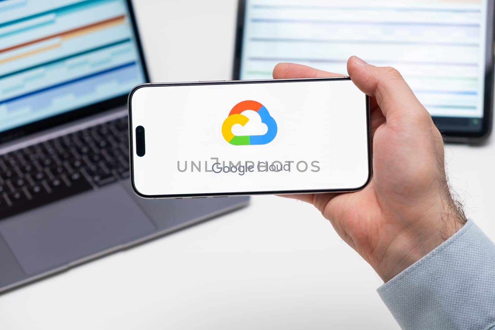 Google Cloud logo of app on the screen of mobile phone held by man in front of the laptop and tablet by vladimka