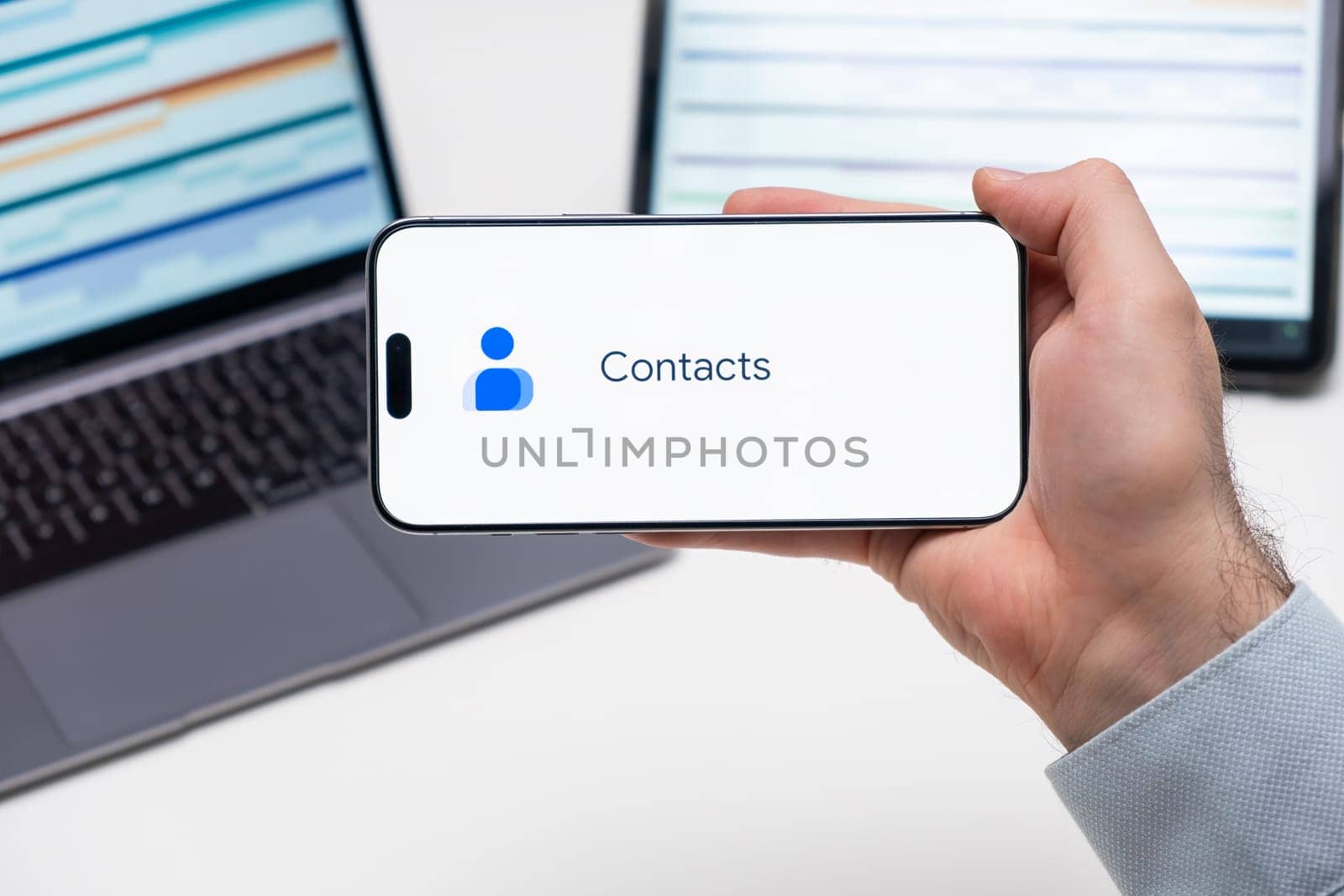Contacts logo of app on the screen of mobile phone held by man in front of the laptop and tablet by vladimka