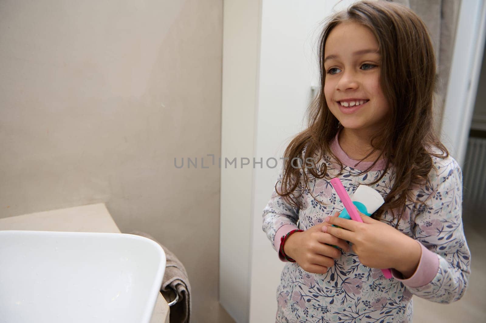 Little child girl in pajamas, holding kids toothpaste and her pink toothbrush, smiles looking at her mirror reflection, standing in a home bathroom before going to bed or when waking up in the morning