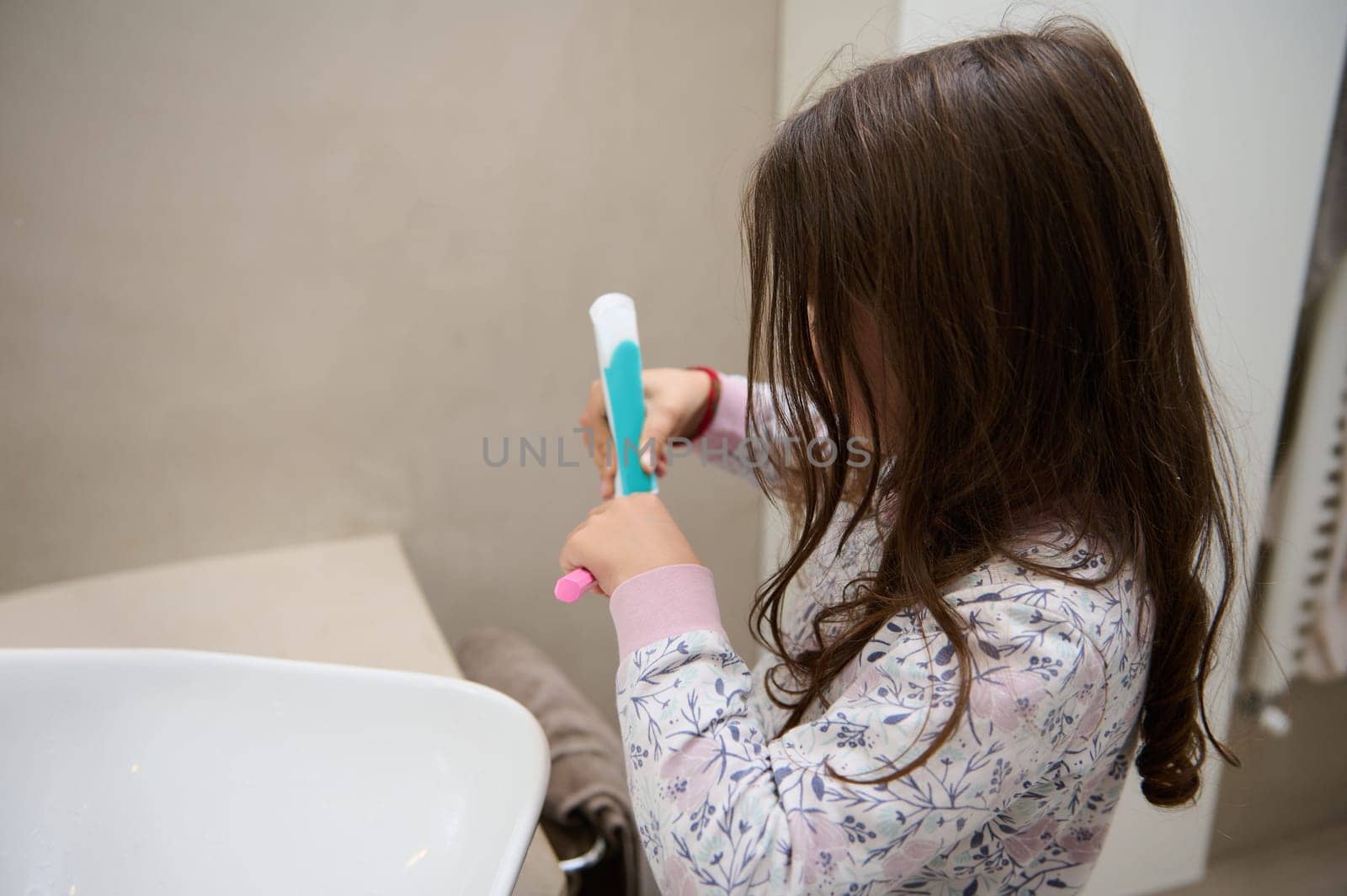 Caucasian little kid girl squeezing toothpaste on her pink toothbrush, standing at white washbasin in the home bathroom by artgf