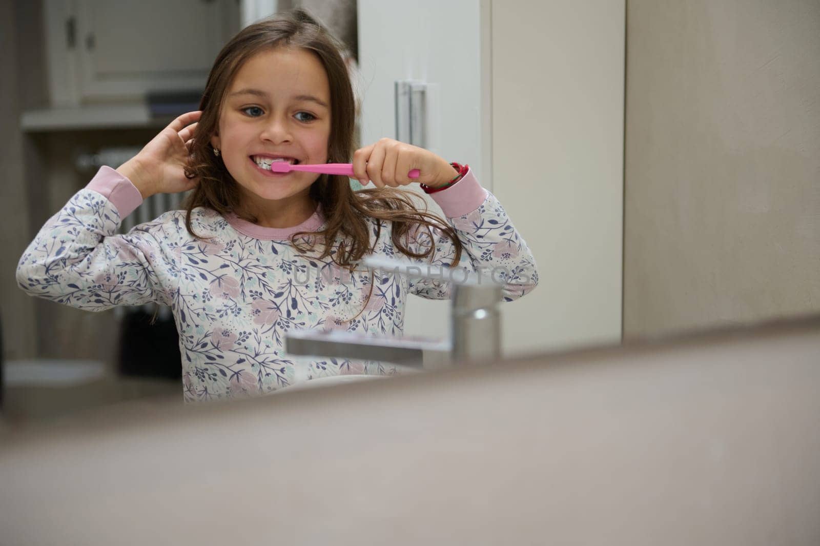 Reflection in the bathroom mirror of a Caucasian cute child girl brushing teeth, dressed in pajamas. Dental hygiene, oral care and healthy lifestyle concept