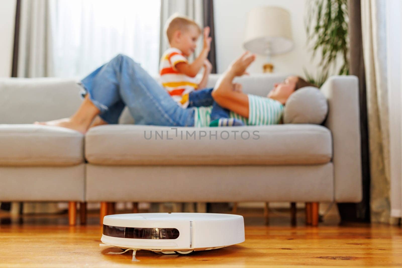 A cheerful mother and her young son playing and relaxing on a couch in a tidy living room with a robotic vacuum cleaner working in the foreground.