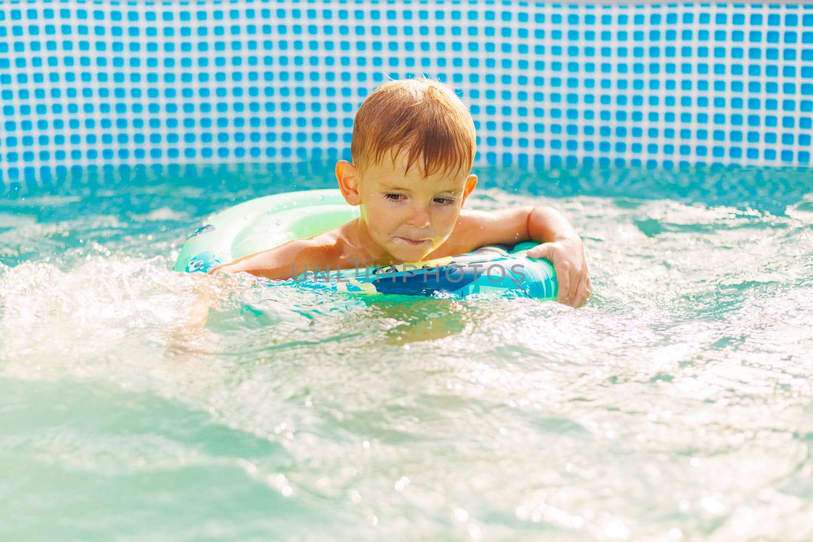 A young boy with a focused expression is learning to swim in a sunny pool using a colorful float ring for safety and support.