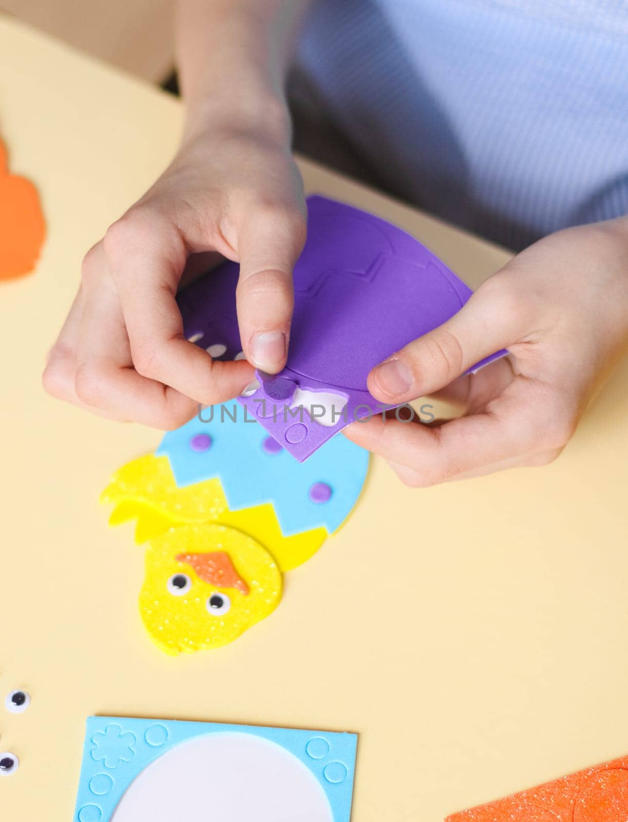 The hands of a caucasian teenage girl peel off a lilac bow sticker with her fingers to stick on a yellow chicken felt, sitting at a children's table with a set of handmade items on a pale yellow background with selective focus, close-up side view. The concept of crafts, diy, needlework, diy, children art, artisanal, Easter preparation,children creative.