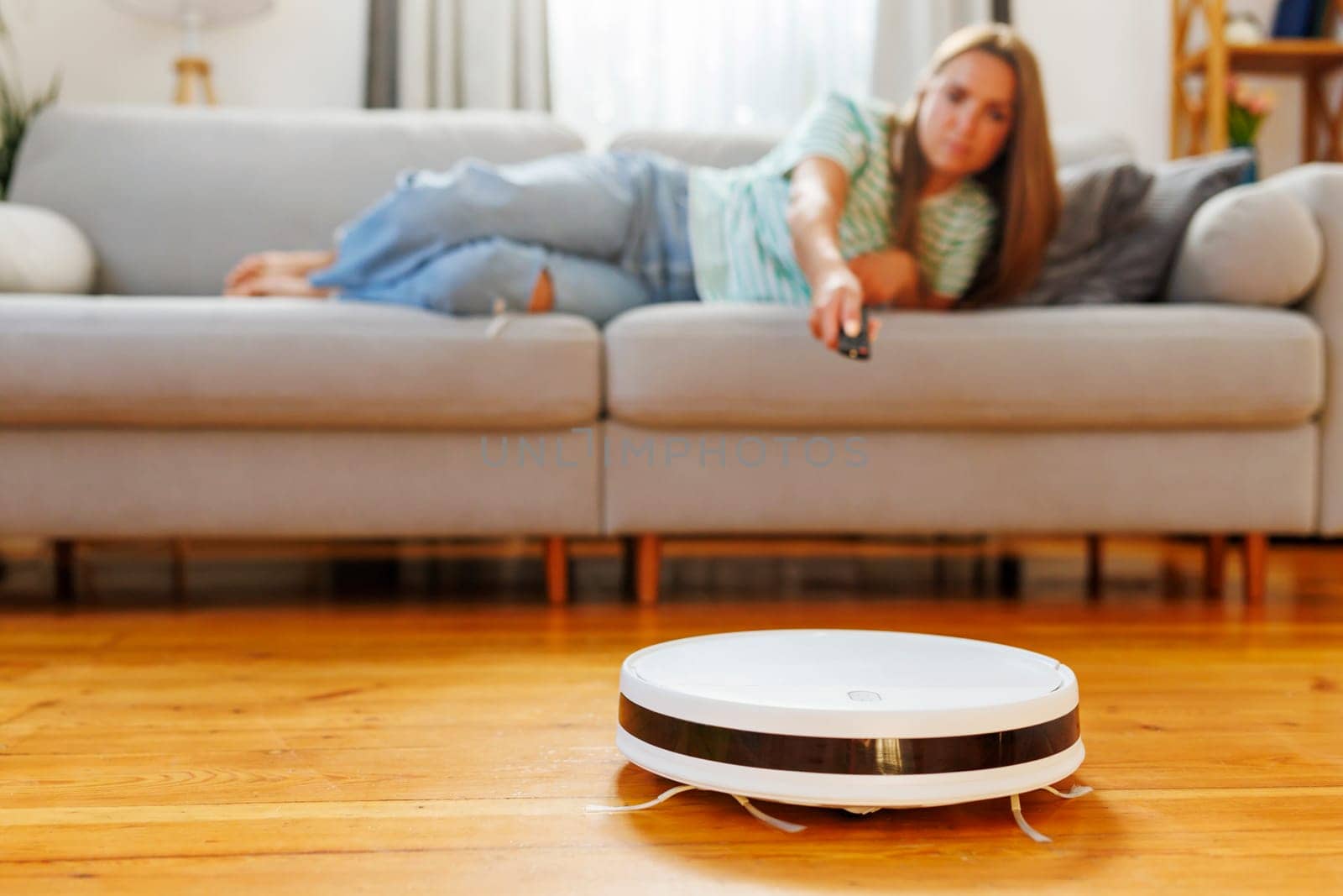 A young woman lounges on the sofa, using a remote control to navigate a robotic vacuum cleaner across the wooden floor of a cozy living room.