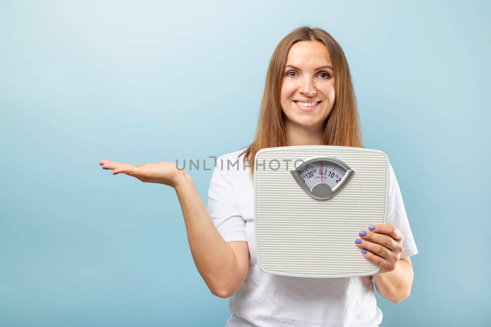 Cheerful woman holding a bathroom scale, making a presenting gesture with her hand against a light blue background.