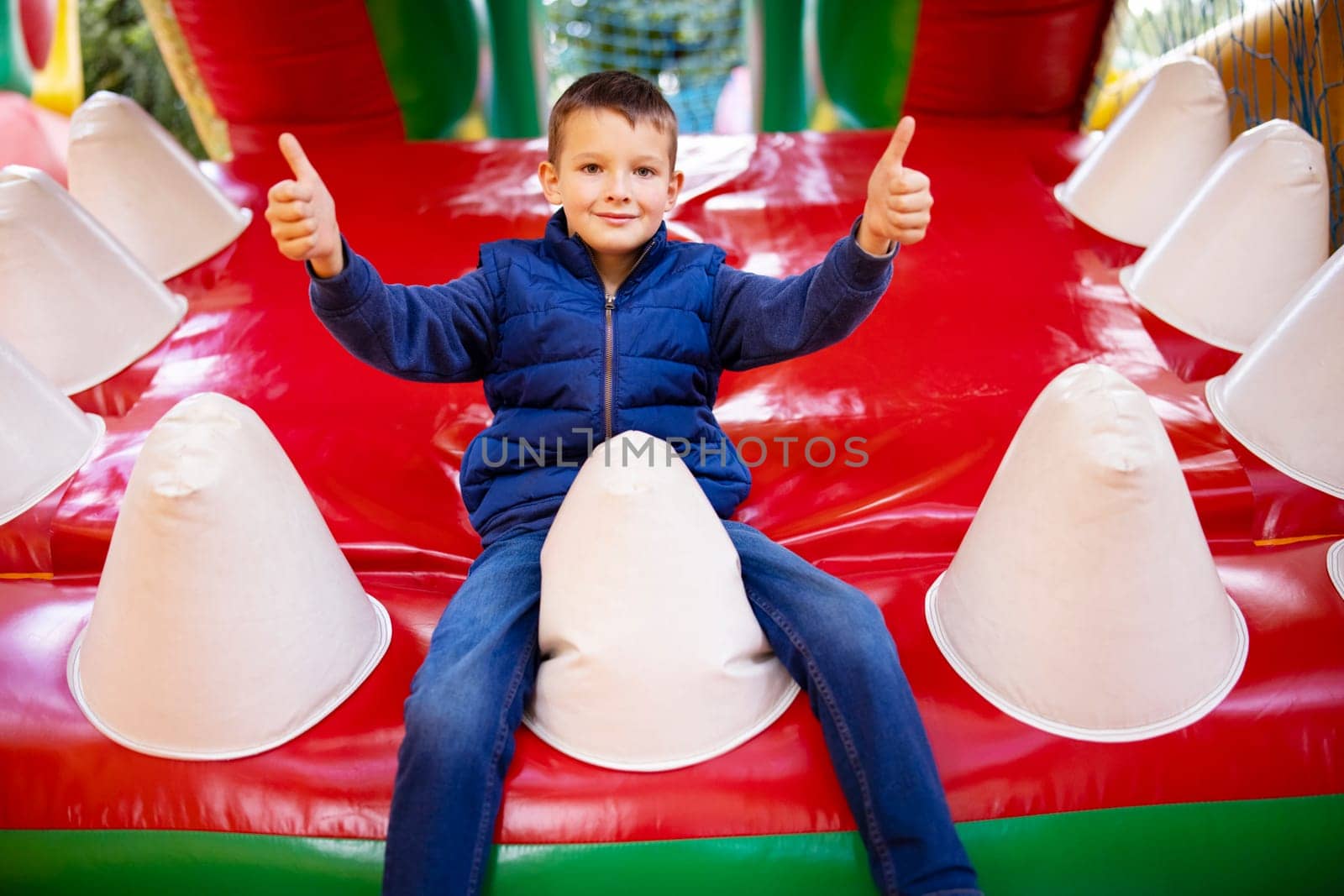 A cheerful young boy in a blue jacket giving two thumbs up while sitting on a colorful inflatable slide at a playground.