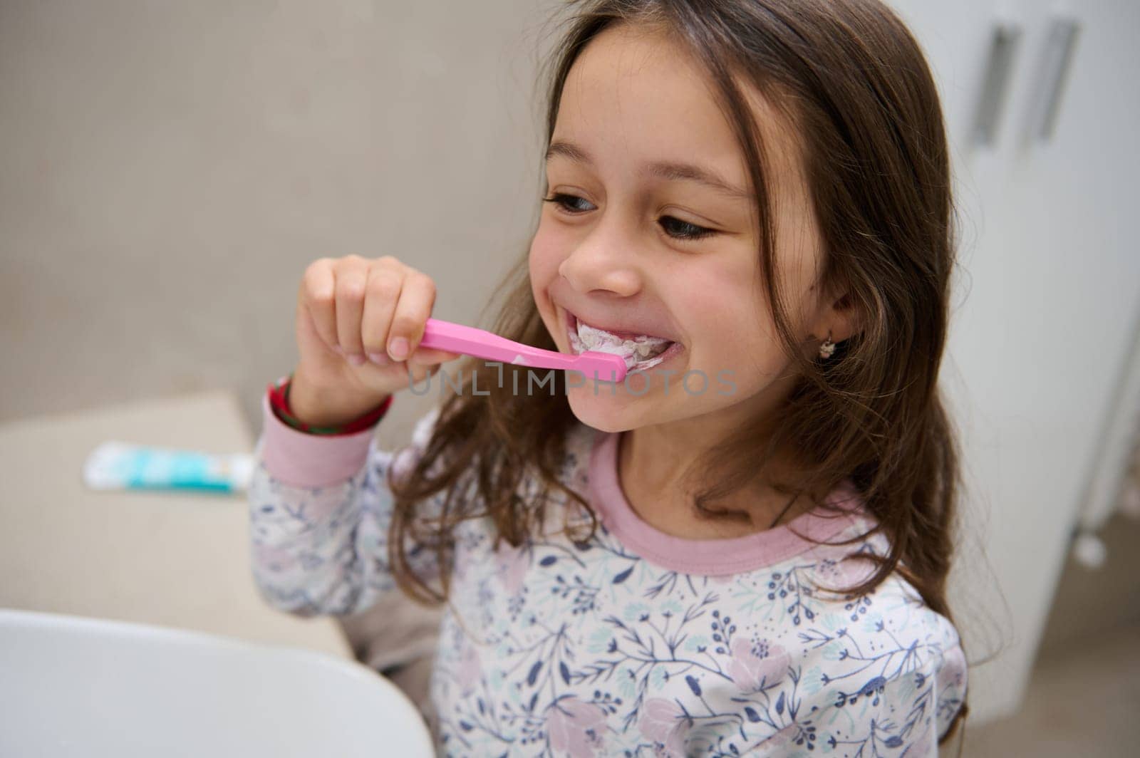 Adorable little child girl in pajamas, holding a toothbrush with toothpaste and brushing her white teeth in front of the mirror in the home bathroom. Healthy lifestyle. Dental oral care and hygiene