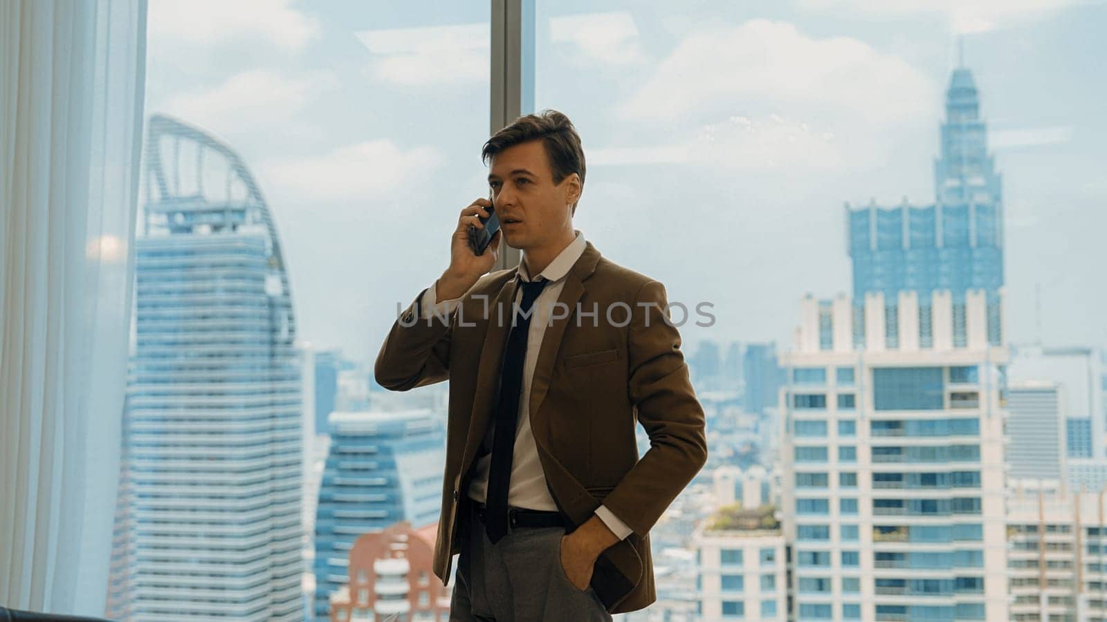 Businessman standing in ornamented office talking on phone with college on cityscape skyline window background. Determination and ambition drive business career toward to bright future