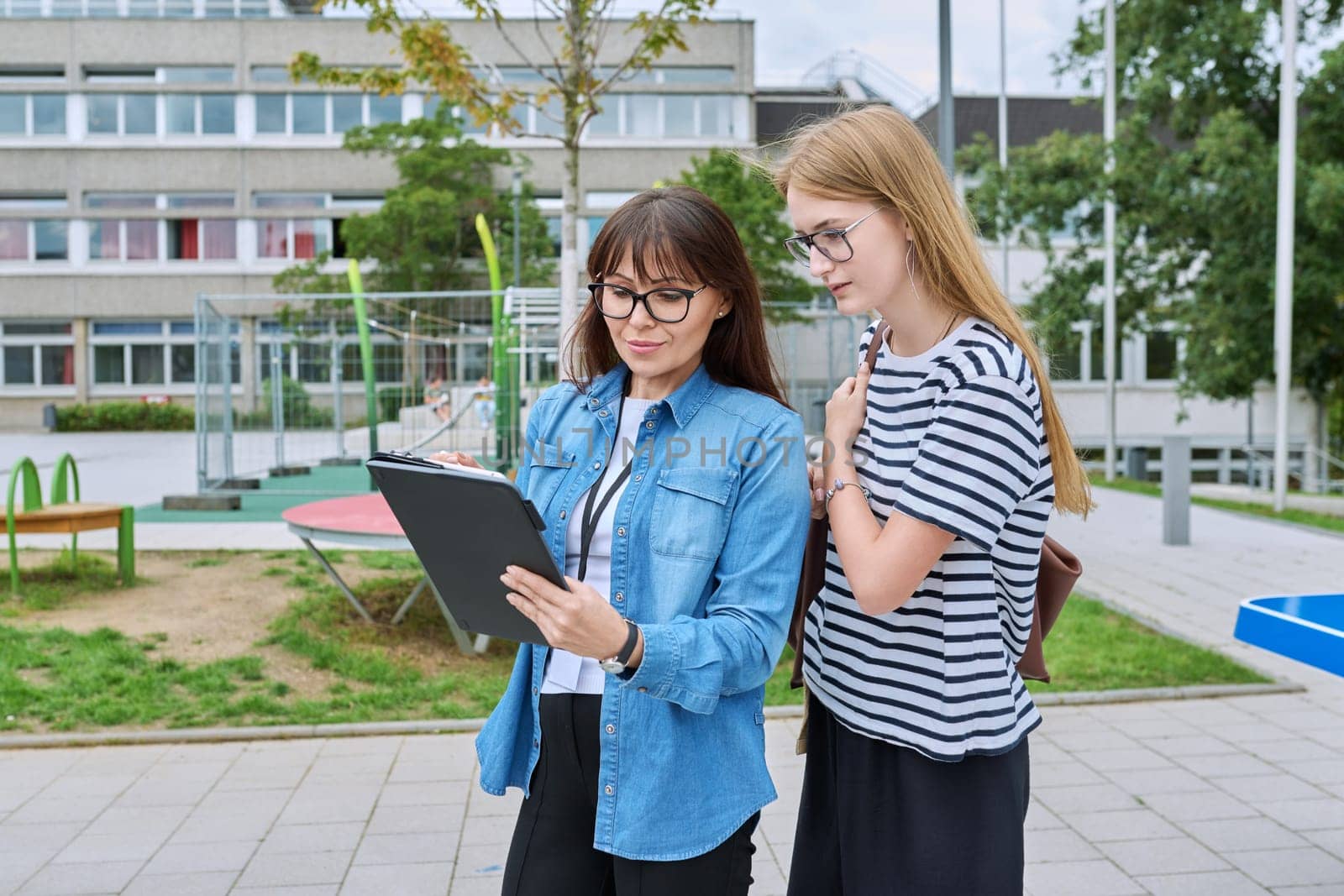 Talking female teacher and teenage high schoolgirl outdoor, school building background. Meeting communication student girl with backpack and mentor counselor. Education, adolescence, learning concept