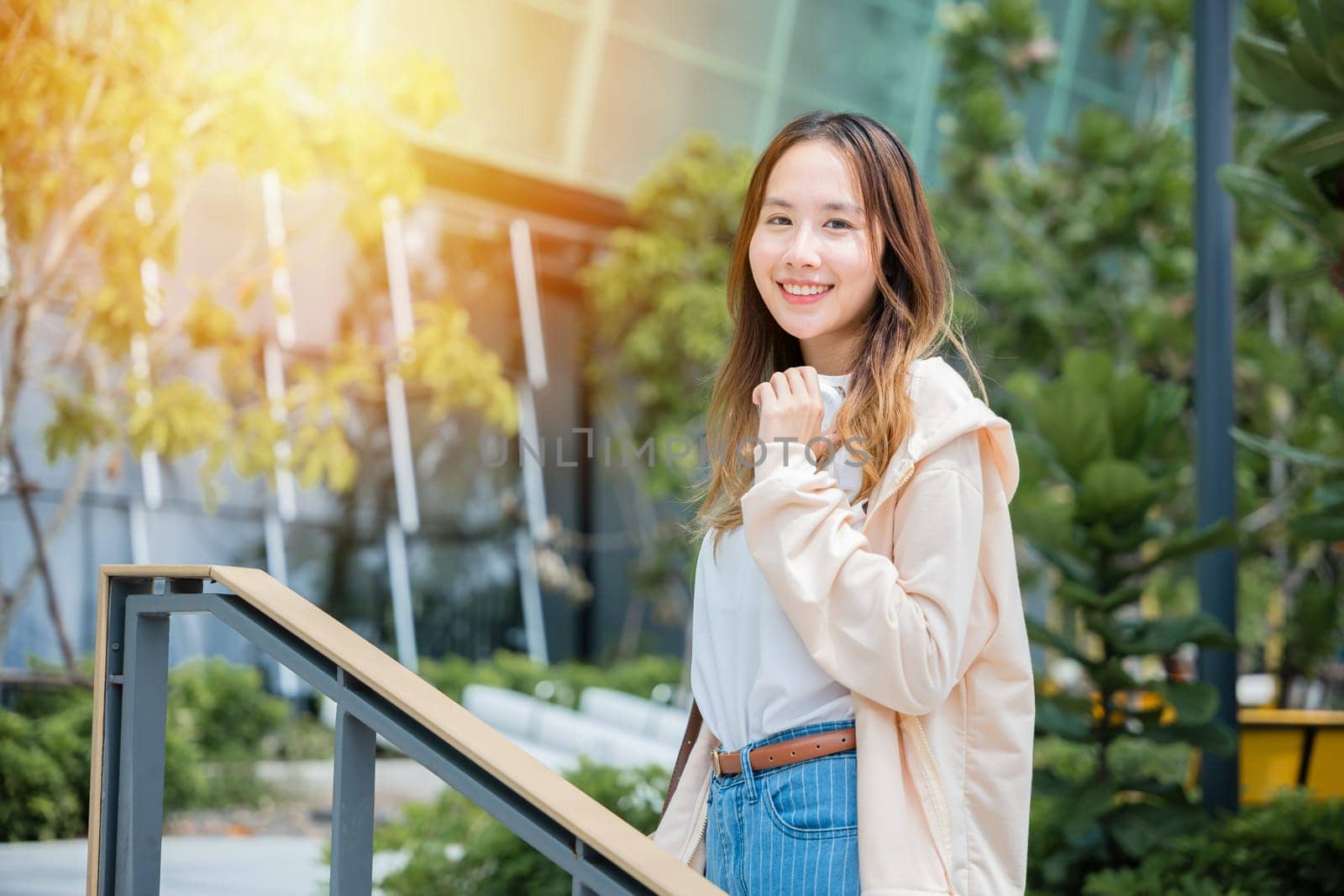 In the city center a smiling Asian woman holds headphones finding joy and connection through music in the heart of the urban landscape.