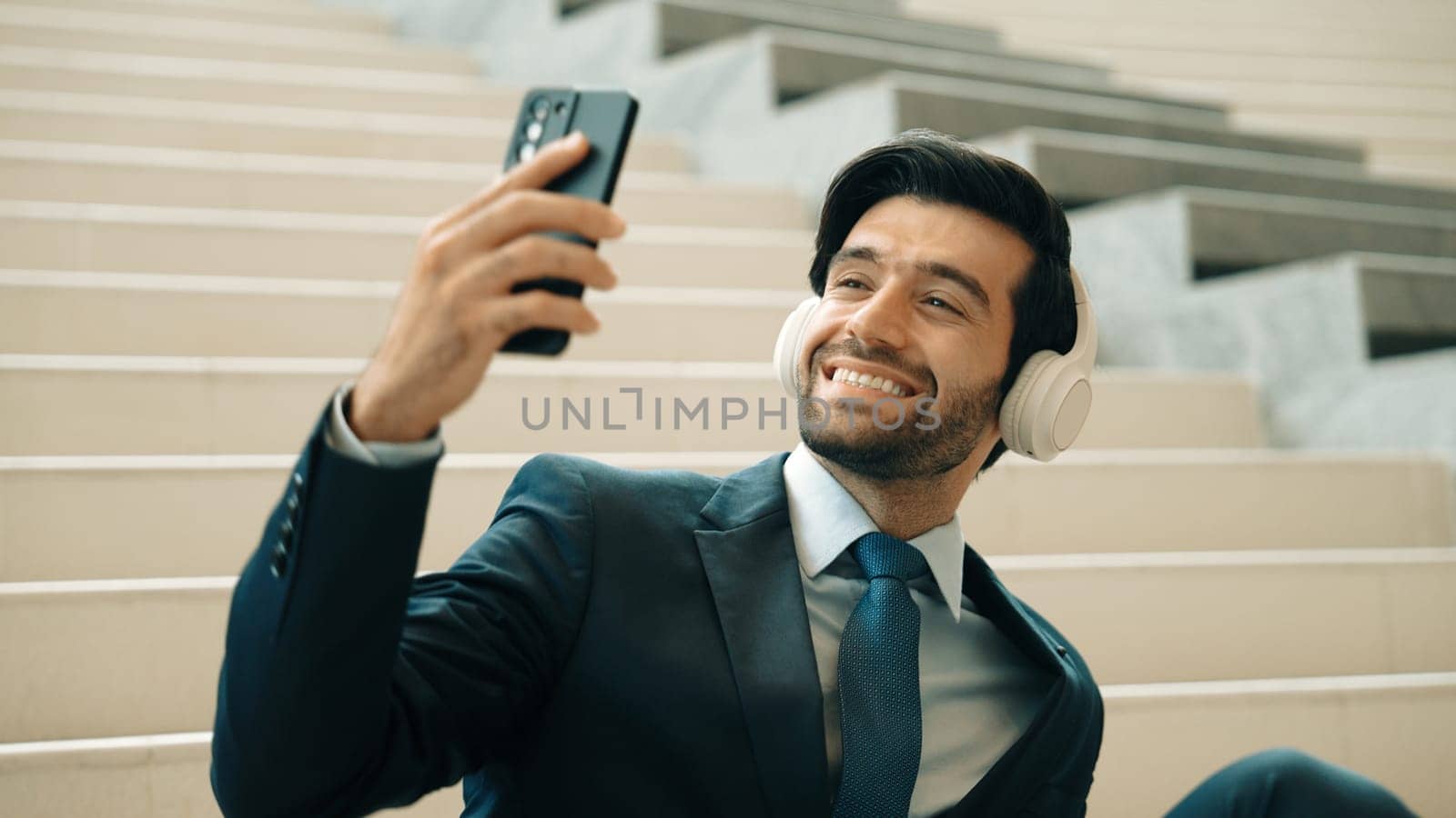 Happy smart business man taking selfie while smiling at smart phone. Closeup image of professional executive manager sitting at stairs while wearing suit and headphone. Creative business. Exultant.