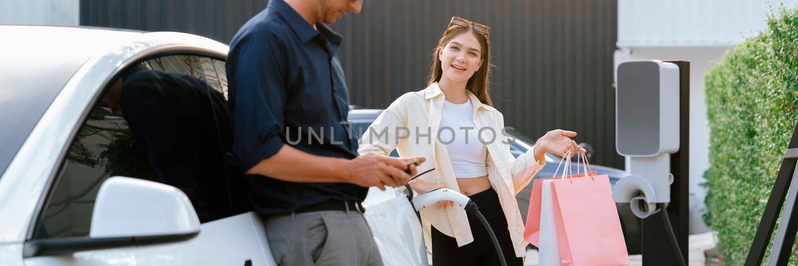 Young couple with shopping bag use smartphone to pay for electricity to recharge EV car battery from charging station at city mall parking lot. Happy couple go shopping by eco car.Panorama Expedient