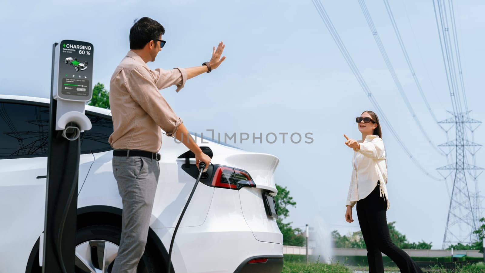 Young couple recharge EV car battery at charging station connected to power grid tower electrical industrial facility as electrical industry for eco friendly vehicle utilization. Expedient
