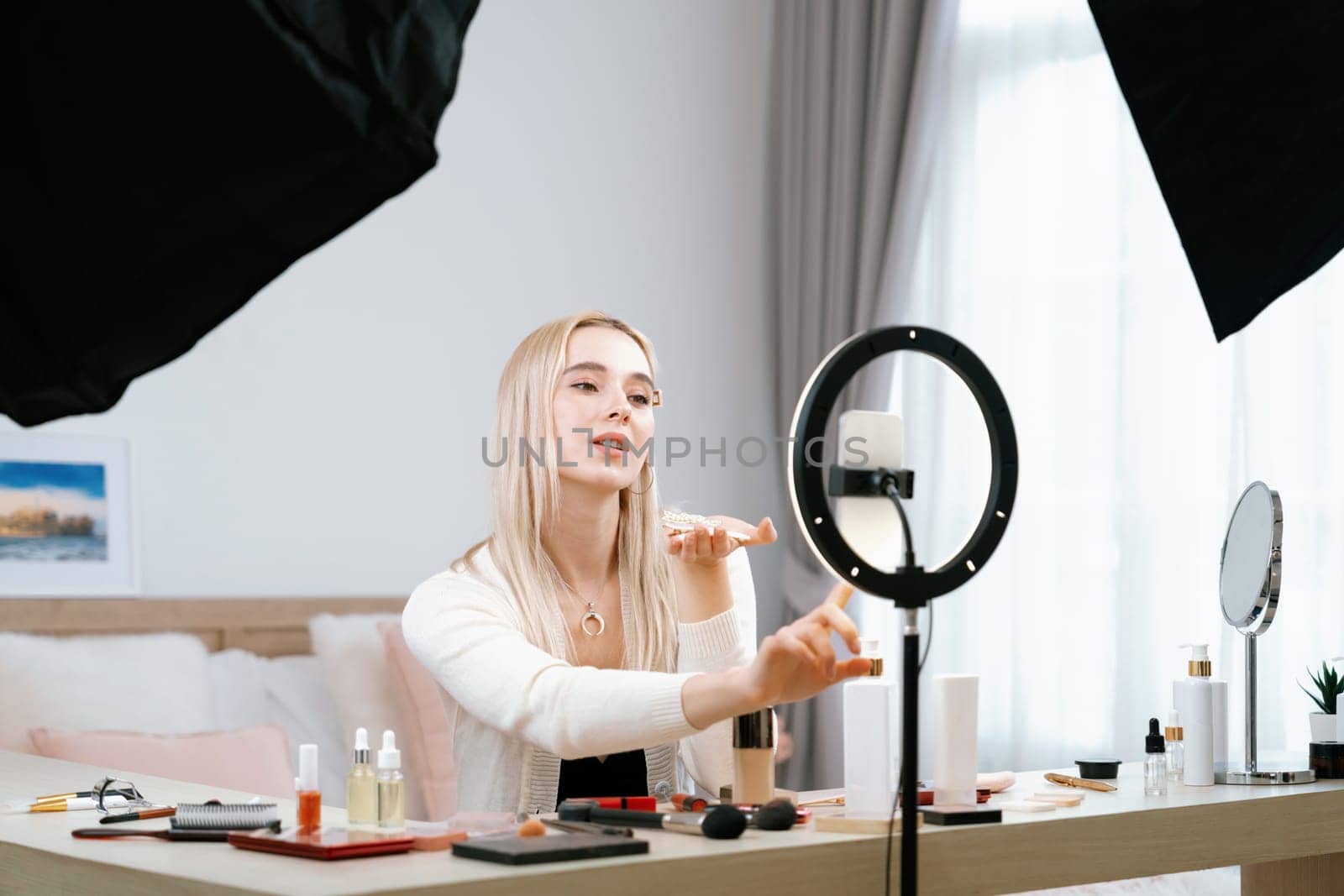 Young woman making beauty and cosmetic tutorial video content for social media. Beauty blogger smiles to camera while showing how to apply beauty skincare to audience or followers. Blithe