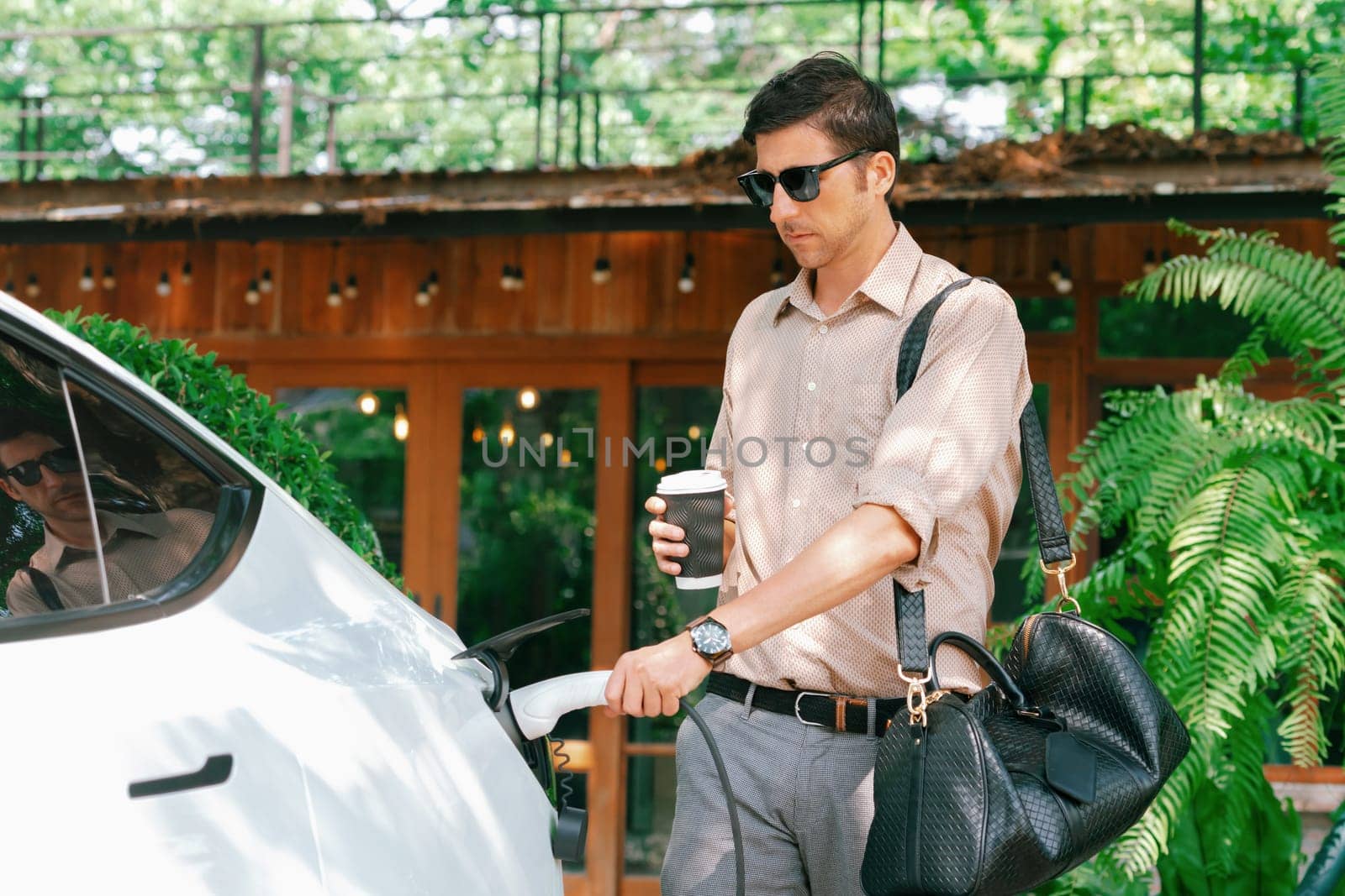 Eco friendly man and sustainable urban commute with EV electric car recharging at outdoor coffee cafe in springtime garden, green city sustainability and environmental friendly EV vehicle. Expedient
