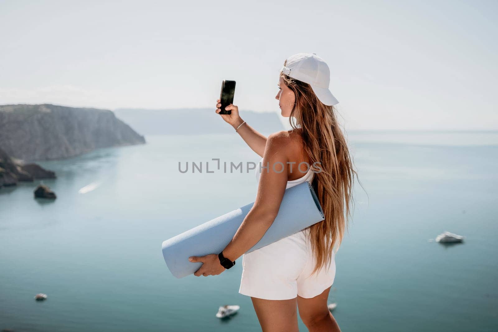 Woman with yoga mat over calm azure sea. Well looking middle aged woman with long hair, fitness instructor with blue yoga mat near the sea. Female fitness yoga routine concept. Healthy lifestyle. by panophotograph