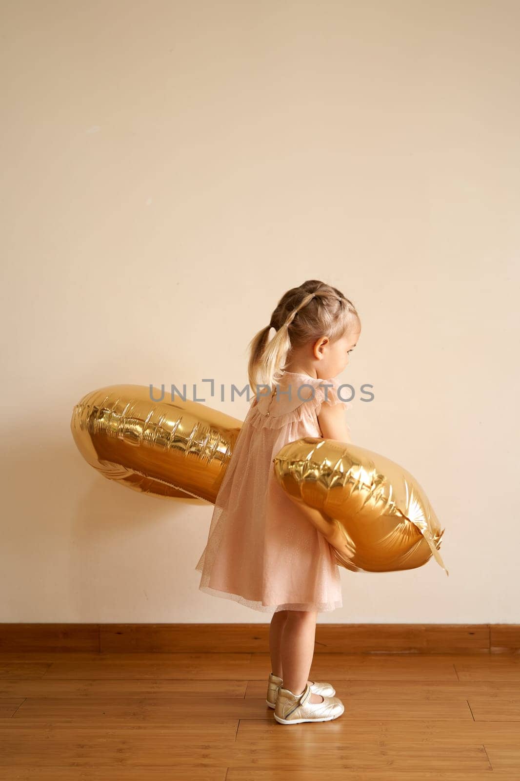 Little girl with an inflatable golden figure in her hands stands against the wall. Side view by Nadtochiy