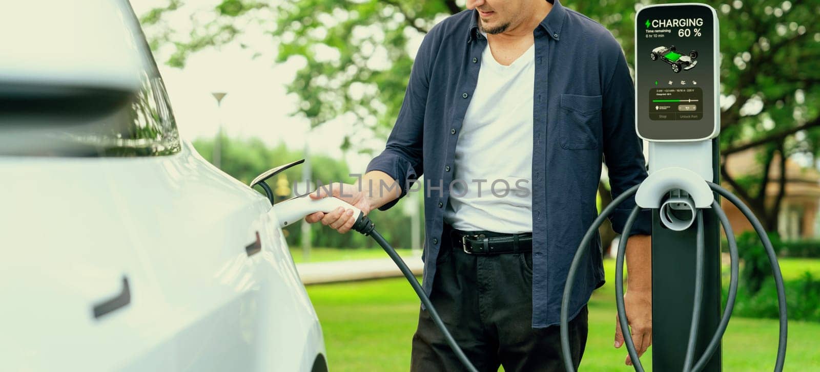 Man recharge EV electric vehicle's battery from EV charging station in outdoor green city park scenic. Eco friendly urban transport and commute with eco friendly EV car travel. Exalt