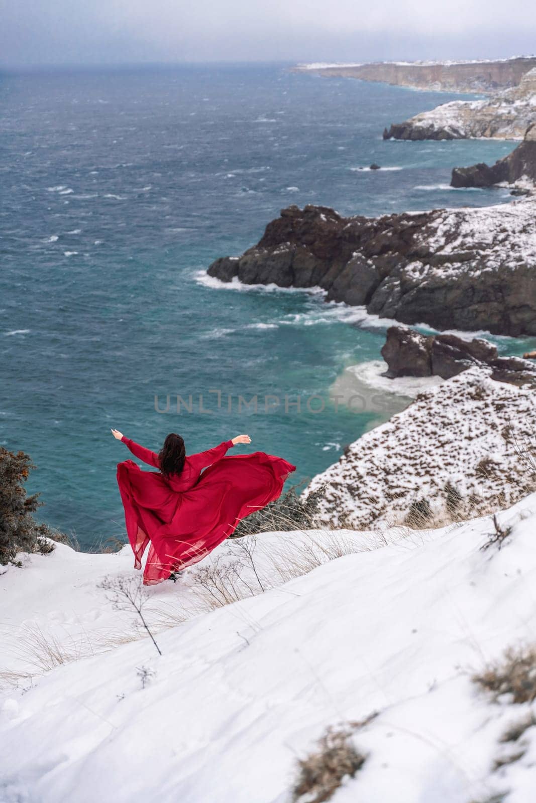 Woman red dress snow sea. Happy woman in a red dress in the snowy mountains by the emerald sea. The wind blows her clothes, posing against sea and snow background