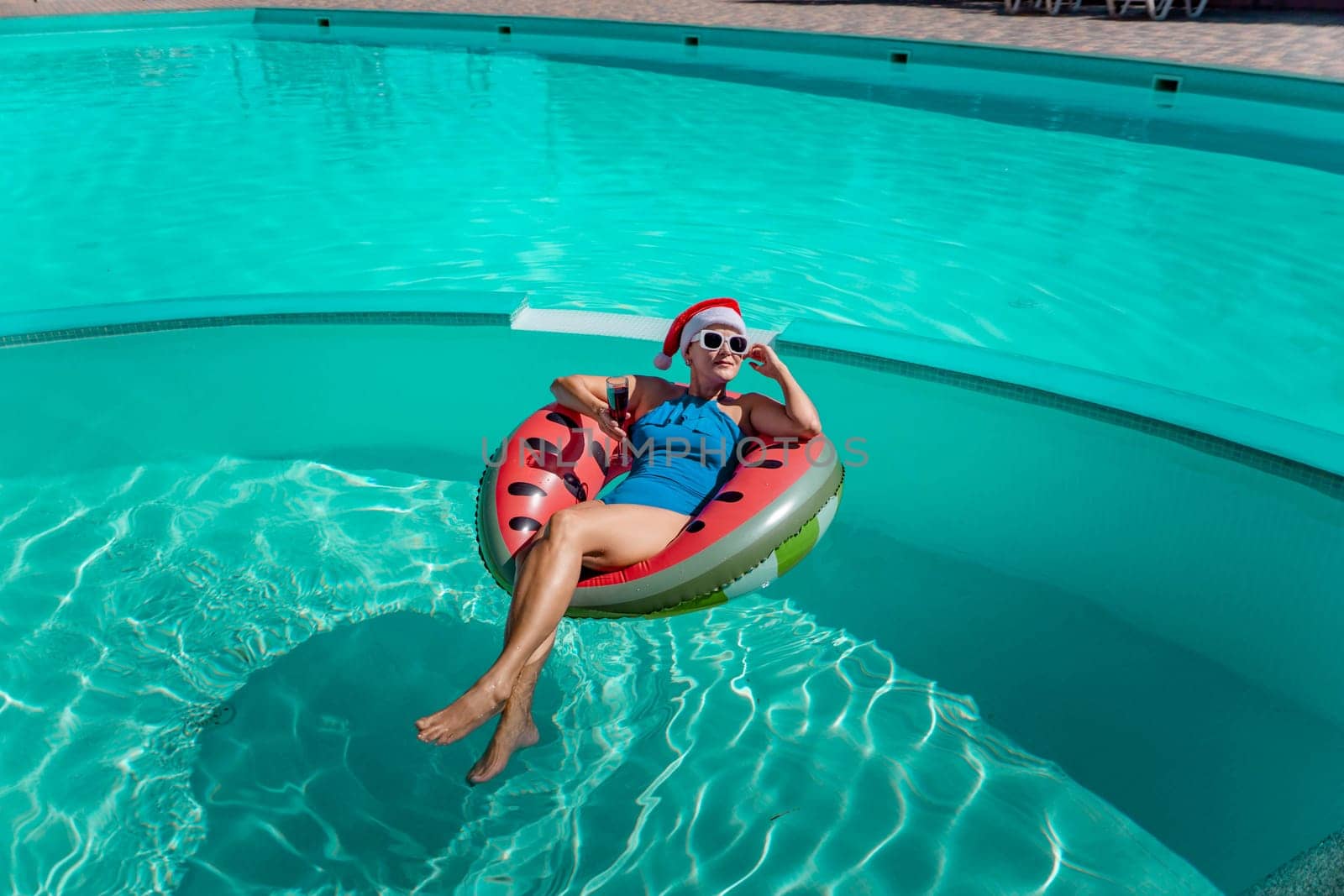 A happy woman in a blue bikini, a red and white Santa hat and sunglasses poses in the pool in an inflatable circle with a watermelon pattern, holding a glass of wine in her hands. Christmas holidays concept. by Matiunina