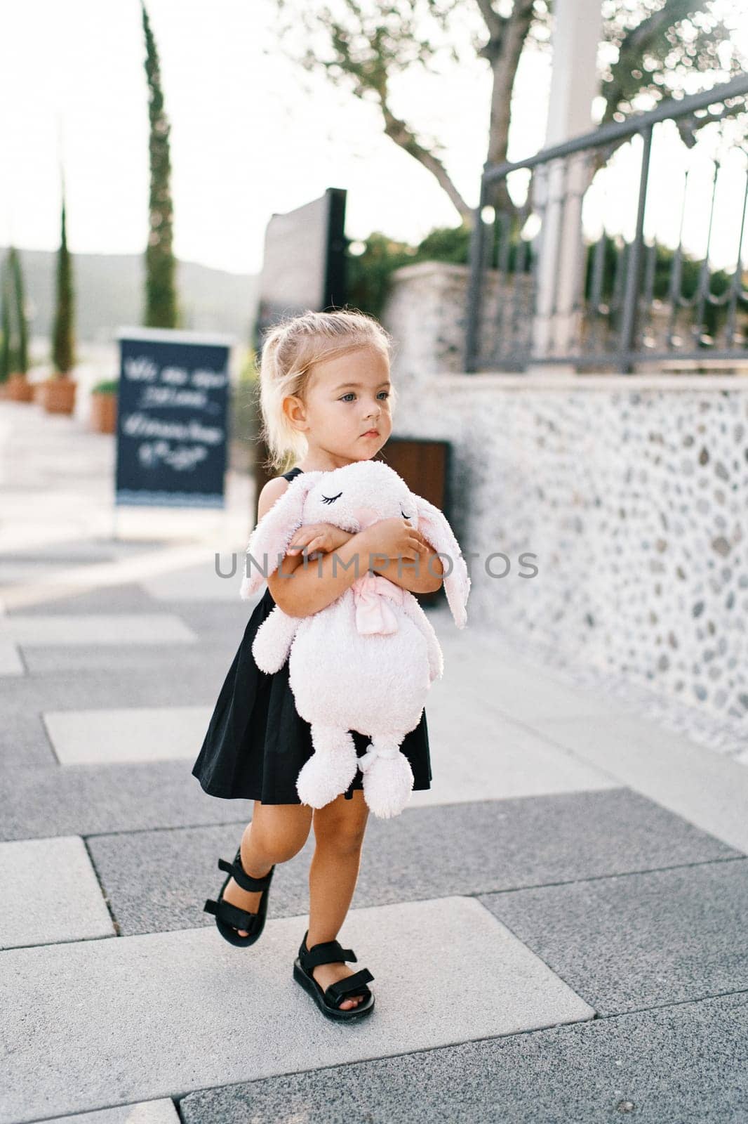 Little girl walks along the road hugging a big soft toy hare. High quality photo