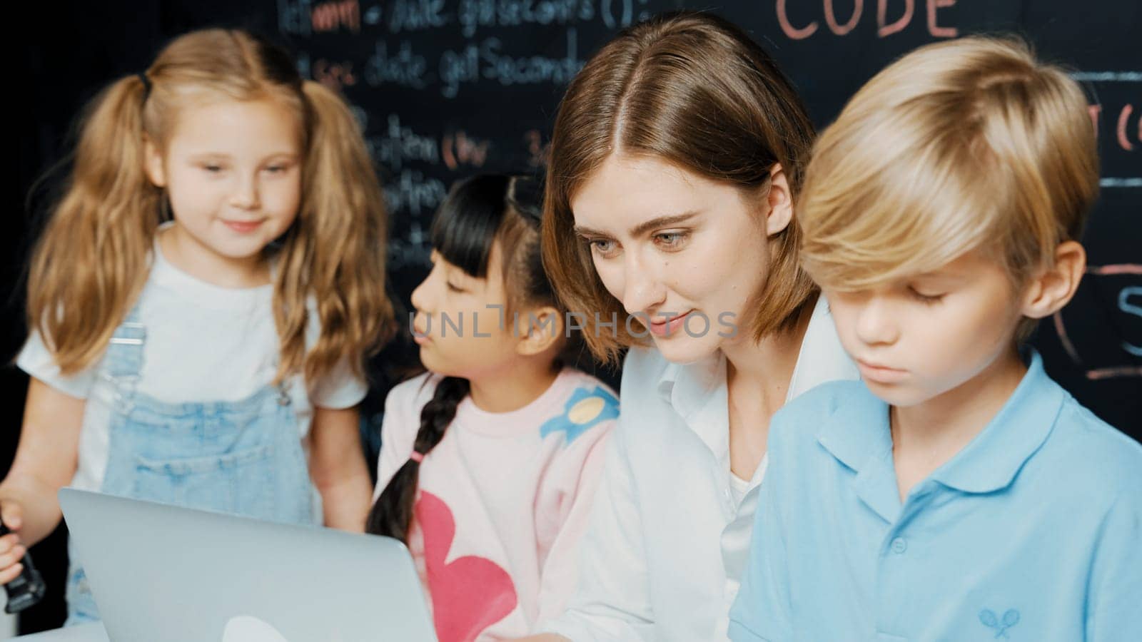 Young smart teacher using laptop to teach diverse student programing system. Caucasian learner looking at computer screen while study about coding prompt at blackboard with program code. Erudition.