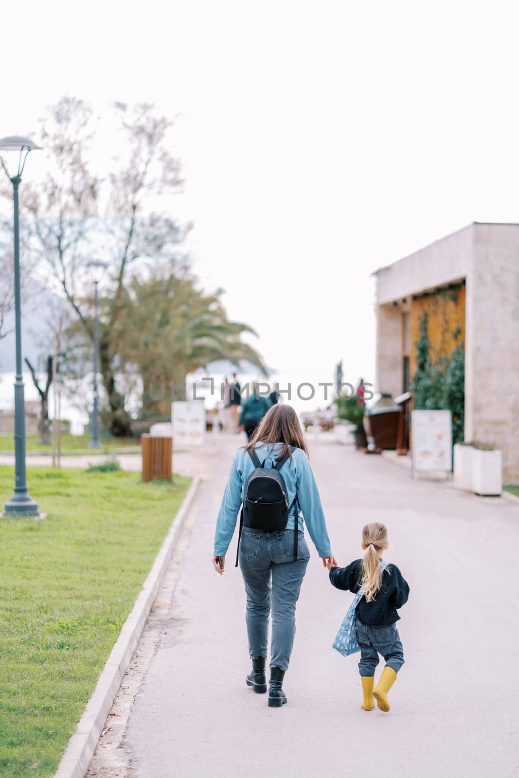 Mother and her little daughter are walking along the road in a green park. Back view by Nadtochiy