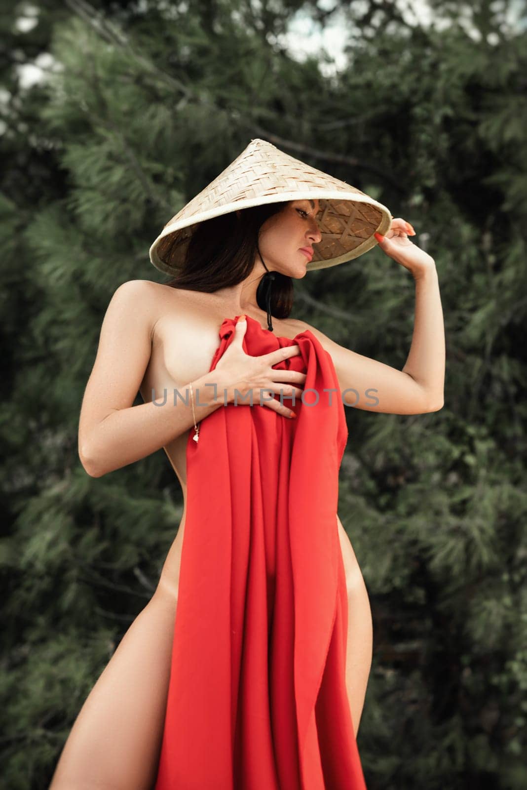 naked sexy woman in red fabric and Asian triangular hat on the background of nature by Rotozey
