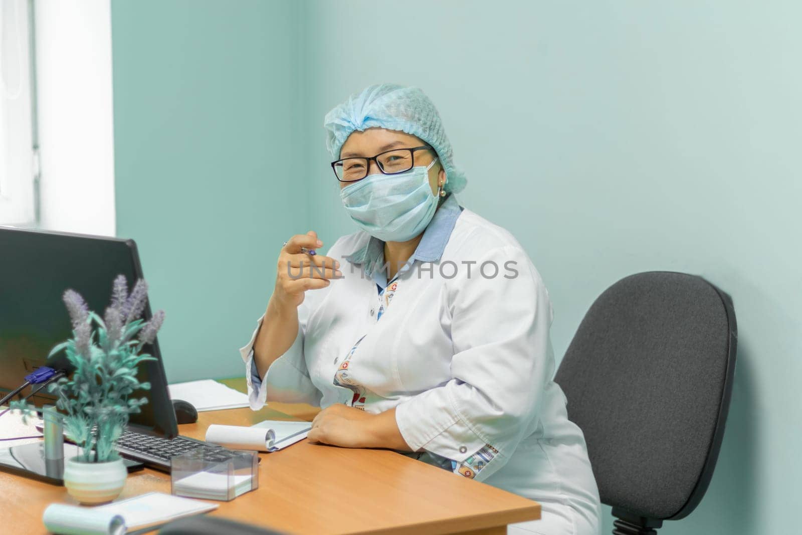 Asian woman doctor waiting for patient. doctor in medical suit, medical cap and mask with glasses, sits in his office at his desk. soft focus by Leoschka