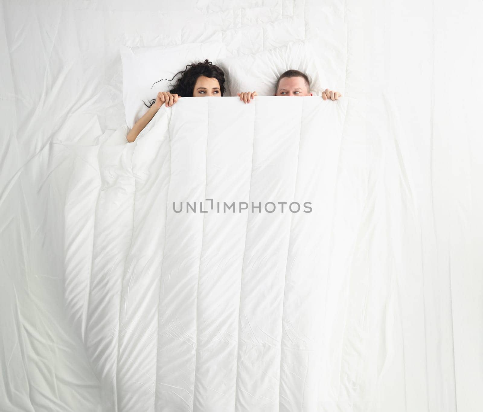 Top view of woman and man peeking on each other under white blanket, awake couple in bed. Good morning, play, fun, family, chilling, sweet home concept