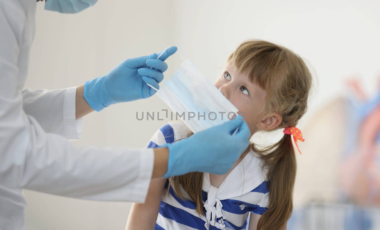 Portrait of doctor put face mask on kid face to protect from virus spread. Little girl sit in hospital and get checkup. Medicine, covid prevention concept