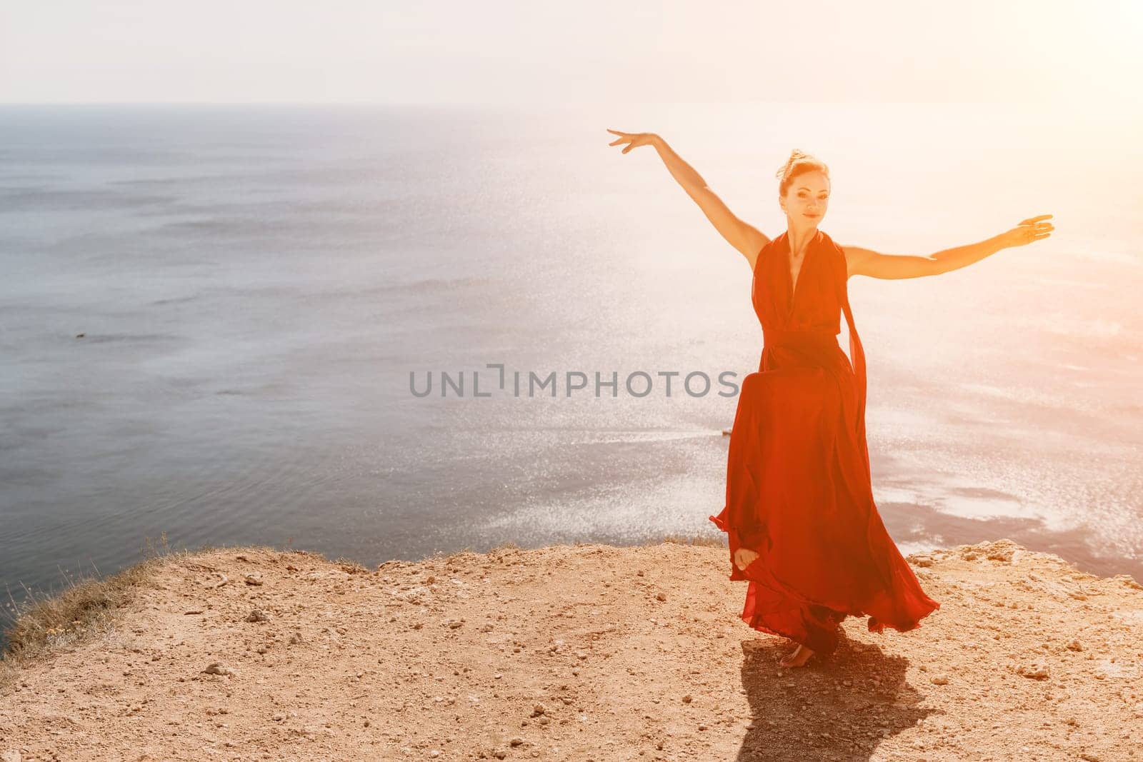 Side view a Young beautiful sensual woman in a red long dress posing on a rock high above the sea during sunrise. Girl on the nature on blue sky background. Fashion photo.